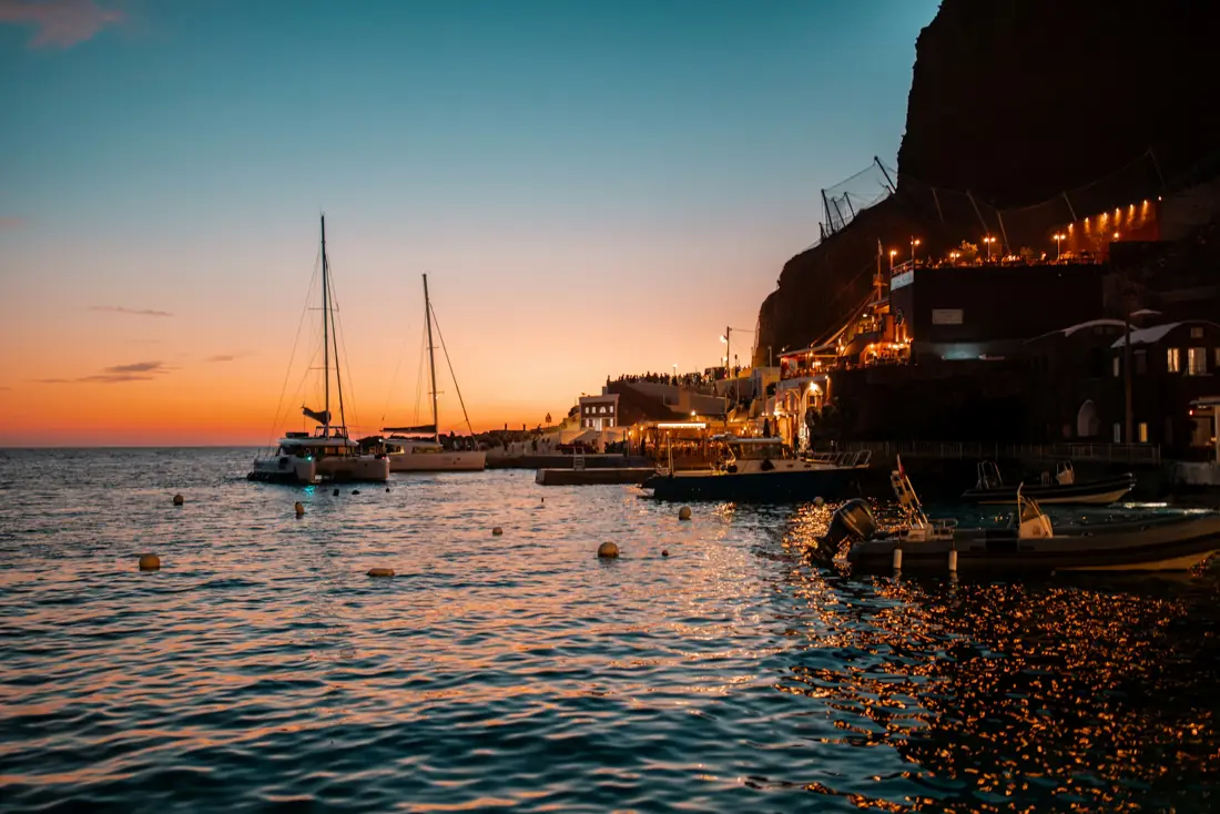 Sailboats in a harbor at sunset beneath lit cliffs on a Greek island
